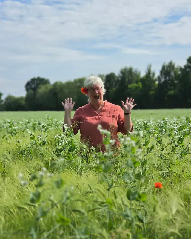 Inken Kuhn steht lachend und winkend mitten in einem Getreidefeld, eine Mohnblume im Haar.