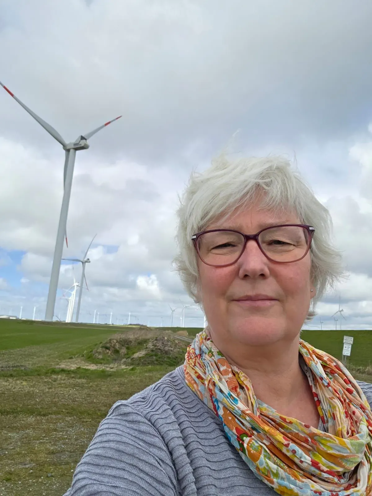 Selfie von Inken Kuhn mit buntem Schal vor Windrädern auf einem Deich.