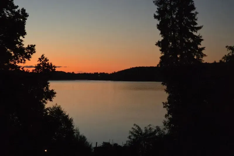 Ein See am Abend, dunkle Bäume am Ufer und ein orange leuchtender Himmel nach Sonnenuntergang.