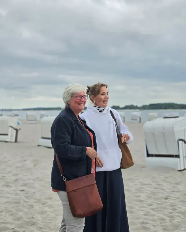 Inken Kuhn und eine Frau stehen lachend am Strand vor Strandkörben.