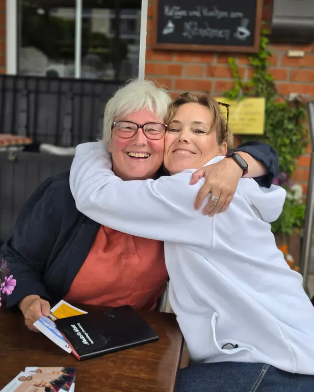 Inken Kuhn und eine Frau umarmen sich lachend an einem Café-Tisch.
